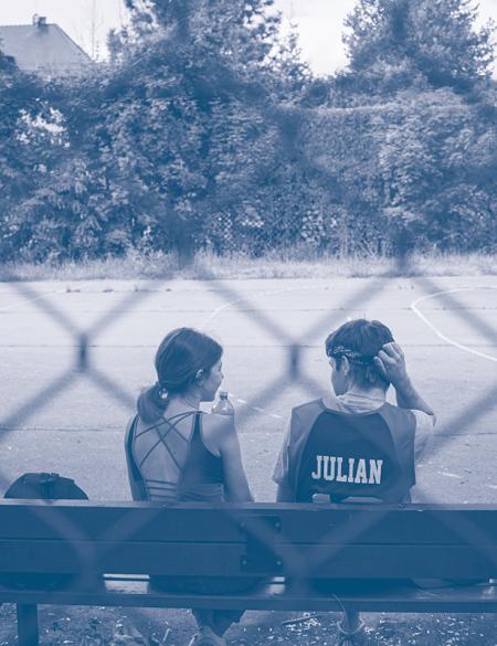 zdjęcie ukazujące dwoje dzieci siedzących na ławce, na boisku. widok zza siatki ogrodzeniowej. 