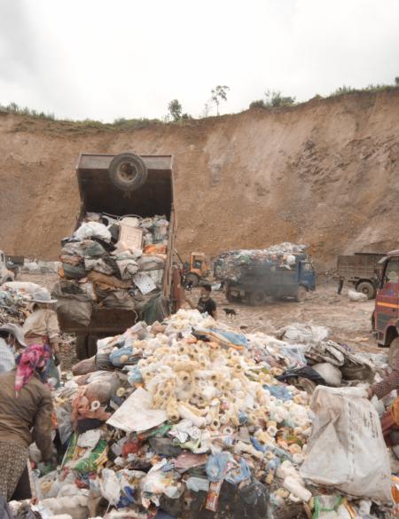 zdjęcie ukazuje ludzi na wysypisku śmieci, w tle ściana ziemi oraz ciężarówki wysypujące odpady